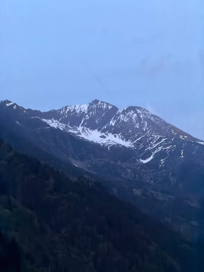 The image displays a serene mountain landscape, captured during what appears to be either dawn or dusk. The scene is dominated by a majestic, dark mountain range, partially covered in snow, occupying the right side of the frame. The mountain features rugged, steep rock faces with significant patches of bright white snow and ice, which highlight its contours and create striking patterns against the dark rock. The lower sections of the mountain are enveloped in deeper shadows, suggesting depth and scale.
To the left, a clear, pale blue sky extends across the frame. The sky is largely devoid of clouds, indicating fair weather, with a soft, diffused light that hints at early morning or late evening. This gentle lighting casts a tranquil atmosphere over the entire scene, emphasizing the natural beauty and grandeur of the mountainous environment.
No people, man-made objects, or visible text are present in the photograph. The overall impression is one of untouched natural wilderness. Given the location context of Piotta, Switzerland, the image likely captures a typical snowy alpine vista in that region.