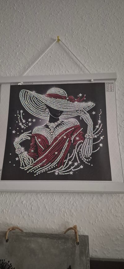 The image shows a diamond painting of a woman wearing a large, ornate hat and a red and white patterned dress. The painting is displayed on a wall, hanging from a yellow thumbtack by white cords. The painting is framed by a white border and appears to be a craft project. A small chart with numbers and symbols is visible in the upper right corner of the painting. Below the painting, a grey object with two rope handles is visible, suggesting it might be part of a decorative display or a craft supply. The wall texture is rough and white, indicative of a textured plaster or wallpaper. The overall scene suggests an indoor setting, possibly a home or studio. There is no discernible information in the image that would allow for the identification of a specific location within Baden-Baden, Germany.