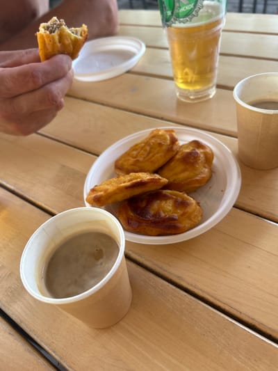 A person's hand holds a partially eaten pastry filled with meat. The pastry is golden brown and appears to be fried. In the background, a white plastic lid sits on a wooden table. To the right of the pastry, a white plastic plate holds four more similar pastries. Next to the plate is a brown paper cup filled with coffee. Further to the right, a clear plastic cup with a green label is filled with a pale yellow beverage, likely beer. The table is made of light-colored wood with visible grain and parallel slats. The scene appears to be outdoors, possibly in a cafe or public eating area. The location is identified as Останинское сельское поселение, Ukraine.