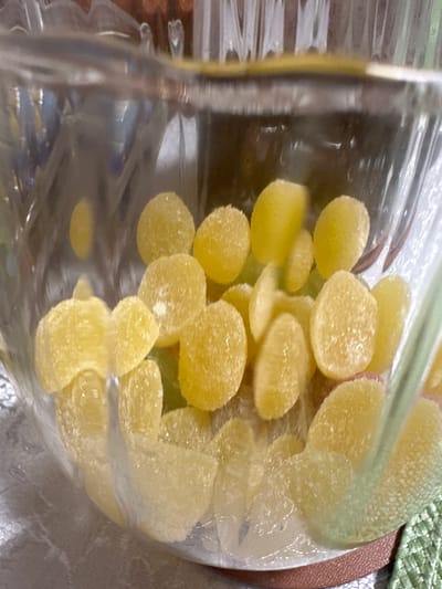 The image depicts a clear, likely plastic, container filled with yellow, round, sugar-coated candies. The container appears partially full, with the candies clustered together. The background shows a blurred surface, potentially a table, with a hint of green and brown. The image is taken indoors with natural lighting. There is no verifiable connection to Vorotynsk, Russia in the image. The description contains no information on people, subjects, actions, interactions, or environment beyond the visual objects present in the image.