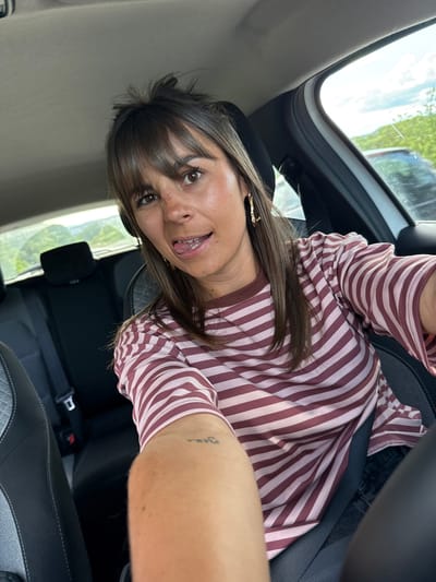 A woman is taking a selfie inside a car. She is wearing a striped shirt and has brown hair with bangs. She is wearing a seatbelt. The car's interior is black, and the seats have a diamond-stitched pattern. The car appears to have a sunroof. The location is Les Arcs, France.