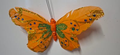 An artificial orange butterfly decoration is displayed against a plain white background. The butterfly has intricate details, including gold glitter accents forming swirling patterns on its wings, and small blue and green rhinestones scattered across them. The body of the butterfly is segmented and appears to be made of a textured, orange material. Two thin black wires, likely for hanging or display, extend from the top of its head. The image is well-lit, with the butterfly clearly in focus. There is no indication of a specific location in Orimattila, Finland, or any interaction with people or animals.