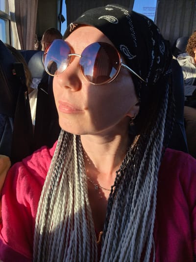 This is a close-up, waist-up, indoor shot of a woman with braided hair and sunglasses, likely taken on a bus. The woman is wearing a pink shirt and a black bandana. Sunlight is visible on her face. She appears to be looking to her left, away from the camera. In the background, there are blurred figures of other passengers and the interior of a bus. The exact location of Verkhniye Tatyshly, Russia, is not discernible from the image itself, but it is presented as the context for this photograph.
