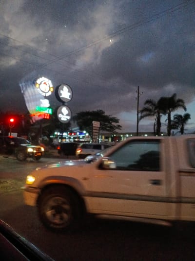 A street scene at dusk or night, taken from a car. Lit store signs are visible across the road. Palm trees stand against the cloudy sky. The white car appears to be a pickup truck. Other vehicles, including motorcycles, are present on the road. A red traffic light is glowing. The image is blurry and rotated.