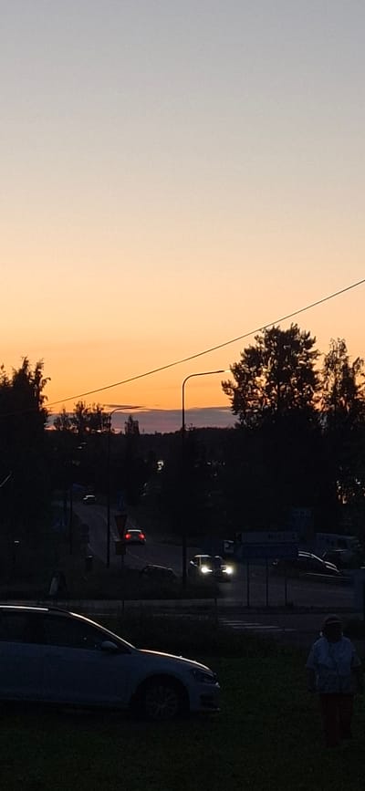 The image depicts a twilight scene in Salpakangas, Finland, characterized by a gradient of soft orange and pale grey in the sky. Silhouetted trees frame the horizon, with a distinct street lamp visible in the middle ground. A road curves through the landscape, with several cars on it. One car's headlights are illuminated, indicating movement. In the foreground, a light-colored car is parked on grassy terrain. A person wearing a light-colored shirt and dark pants is standing to the right of this foreground car. The overall impression is a serene, transitional moment between day and night in a suburban or rural setting.