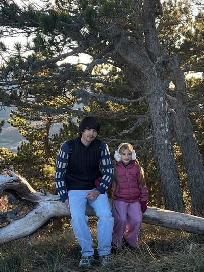 A man and a girl lie on a fallen tree trunk in a forest. The man wears a black vest over a sweater with a pattern, light blue jeans, and sneakers. The girl is dressed in a burgundy jacket, lavender pants, and white earmuffs. They are resting against the trunk, with evergreen trees and a valley visible in the background. Sunlight filters through the branches above.
