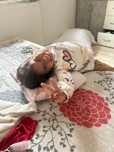 The image shows a young girl lying on a bed in a bedroom. She is smiling and making a peace sign with her fingers. She is wearing pajamas with a heart and star pattern. The bed is covered with a gray and white patterned comforter. There are clothes on the bed next to the girl, including a white fluffy blanket and a red garment. In the background, there is a radiator and a wardrobe with drawers. The overall environment suggests a domestic setting. There is no indication of the location being in Vohburg an der Donau, Germany.