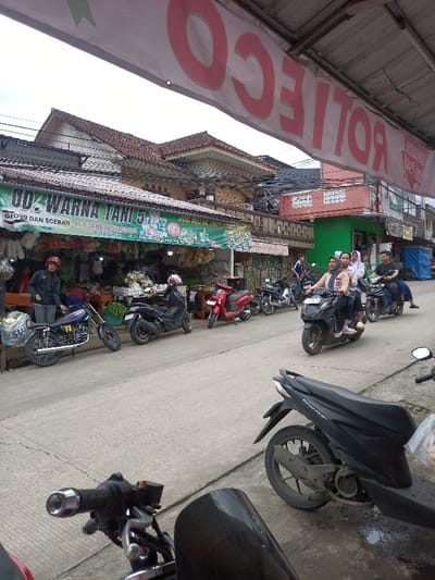 The image depicts a street scene in Caringin, Indonesia, under an overcast sky, viewed from a slightly elevated position behind a parked black motorcycle. In the immediate foreground, the handlebars and a partial black windscreen of the camera-side motorcycle are visible on the left, while a black Honda Beat scooter is parked on the right.
The midground features a concrete road, with multiple motorcycles and scooters parked along the left side. A man wearing a black jacket and a red helmet stands next to a dark blue and gold motorcycle parked closest to the camera on the left. Further down, a black Yamaha NMAX type scooter and a red scooter are parked. On the road, traffic moves from right to left. A black scooter carries three individuals: a male driver and two female passengers, one wearing a white headscarf. Following them, a blue scooter is driven by a male with a male passenger. Another male on a motorcycle is visible further back.
The background on the left reveals an open-front shop with a prominent green banner displaying white and yellow text, including "UD. WARNA TANI 5" and "GROSIR DAN ECERAN JUMLAH KEBUTUHAN." The shop appears to sell produce such as onions and other packaged goods, visible on display tables. Above this shop and further along the street are multi-story buildings with brown tiled roofs and varied architectural styles, suggesting a commercial and residential mix. A large white banner with red, inverted text reading "ROTI EGO" is stretched across the top of the frame. Power lines and utility poles are visible above the buildings.