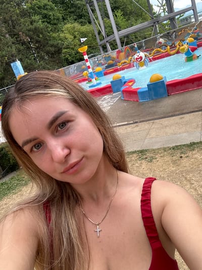 The image shows a young woman taking a selfie in front of a water park attraction. She has long blonde hair and is wearing a red bikini top and a gold necklace with a cross pendant. The water park features a shallow pool with several colorful decorations, including a clown spraying water and a duck-shaped slide. The background includes trees and a metal structure, possibly part of the water park's infrastructure. The scene appears to be outdoors on a sunny day. The location is likely an outdoor water park in Renens, Switzerland, given the contextual information provided.