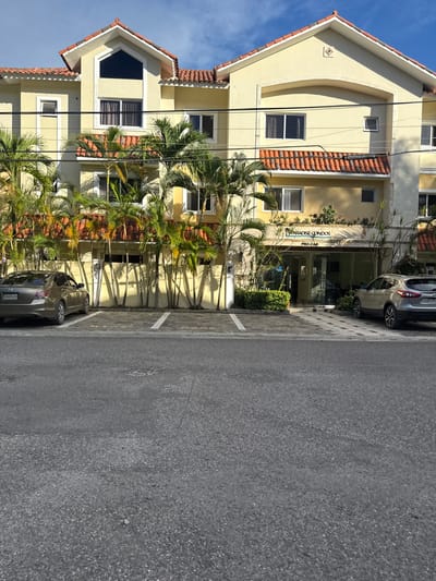 A bright, sunny, daytime outdoor scene in Cabarete, Dominican Republic, depicting a multi-story building and an adjacent asphalt parking lot. The building features a light yellow facade, multiple windows, and distinctive terracotta-tiled roofs and eaves, characteristic of architecture in a warm climate. Numerous palm trees are planted along the front of the building, casting long, sharp shadows onto its walls and the ground, indicating a high sun position. The sky is clear and blue with minimal cloud cover.
In the foreground, an asphalt parking lot with white painted lines is visible. Two cars are parked: a silver SUV near the building's main entrance and a dark sedan parked further down the lot. The license plate of the dark sedan is partially visible, reading "X602619", and a partial plate "G 4157" is seen on the silver SUV.
Above the building's main entrance, a sign is prominently displayed, reading "PARADISE CONDOS" in blue lettering, with "PRO-CAB" visible underneath. No people are visible, and there is no overt activity taking place, conveying a tranquil and static atmosphere. The scene captures a typical view of a residential or tourist accommodation complex in a tropical setting.