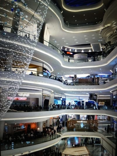 The image shows a multi-level interior of a modern shopping mall. A large, ornate chandelier made of lights and glass beads hangs prominently on the left side of the frame, extending from the top down to the lower levels. Multiple floors are visible, with curved balconies and walkways overlooking a central atrium. Each level features various shops and businesses, indicated by illuminated signage and displays. People are seen walking on the different levels, shopping, and congregating. The architecture is sleek and contemporary, with smooth, curved surfaces and integrated lighting that creates a bright and airy atmosphere. The overall impression is of a bustling commercial space.