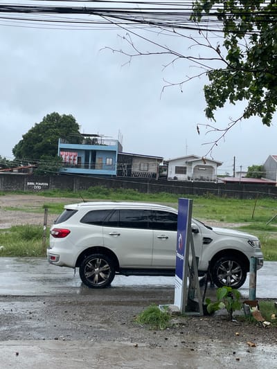 In the city of San Jose del Monte, Philippines, on a wet, overcast day, a white Ford Everest SUV is parked on a wet, paved road adjacent to a grassy area and a concrete wall. The wall has text painted on it that reads "BAWAL PUMARADA DITO". Behind the wall, there are several buildings, including a blue two-story house with a balcony and a white single-story house. Power lines crisscross the sky above, and a tree with green leaves and bare branches is visible in the foreground, obscuring part of the sky. A blue sign with a logo is positioned in front of the SUV. The ground is covered in puddles and debris, indicating recent rain.