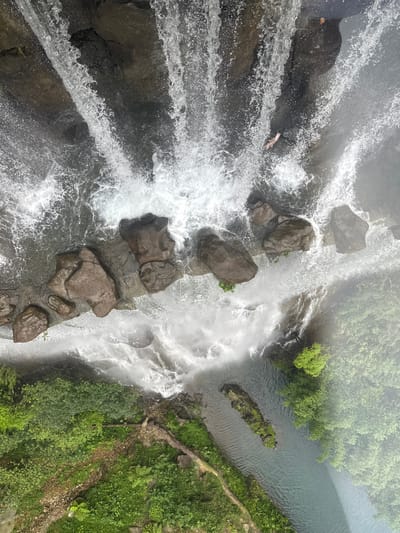 The image is a high-angle, top-down view of a waterfall cascading over rocks into a river. The water is white and frothy, with some of the rocks visible beneath the surface. The riverbanks are lined with lush green vegetation, including trees and bushes. The scene appears to be in a natural, outdoor environment, likely a park or nature reserve. The waterfall is located in Qingyuan City, China. There are no people visible in the image. The image is cropped from the original to provide closer views of different sections of the waterfall and its surroundings.