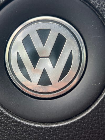 This is a close-up, eye-level, color photograph of a Volkswagen emblem on a steering wheel. The emblem is centrally located within a black, textured circular area. The emblem itself is circular with a silver, metallic finish and features the characteristic VW logo, formed by a 'V' positioned above a 'W'. The logo is inset into the emblem, with the background of the 'V' and 'W' appearing in black. The surrounding circular border of the emblem has a slightly worn, brushed metal appearance with some subtle scratches visible. The black area around the emblem is textured, suggesting the material of a car's interior, likely a steering wheel or dashboard. The lighting appears to be natural and somewhat diffused, creating soft highlights on the metallic surfaces of the emblem. There is no text visible in the image. The context provided indicates the location is Ruzhyn, Ukraine, suggesting this is likely a close-up of a vehicle interior in that region. The image captures a common object found in automobiles, focusing on the brand's insignia.
