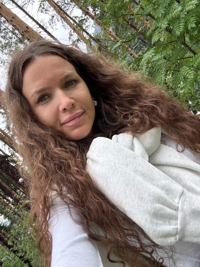 A selfie is taken by a woman with long, wavy brown hair and green eyes, looking towards the camera. She is wearing a white ribbed top and a light gray hooded sweatshirt. She has light makeup on, including lipstick and eyeliner. She is positioned outdoors with trees and foliage in the background, suggesting a natural or park setting. The lighting appears to be overcast.
