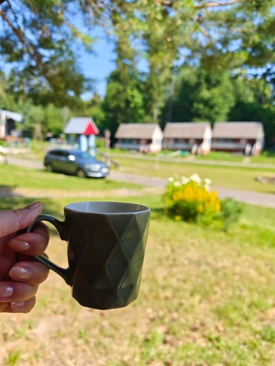 A hand holds a dark green mug with a textured surface in the foreground of the image. The background is blurred and depicts a sunny outdoor scene with green grass, trees, and several small white buildings with light brown roofs. A gray car is parked on a path in the middle ground. There is a playground structure with red and blue accents in the distance. The image appears to be taken at a resort or campsite in Sosny, Ukraine.