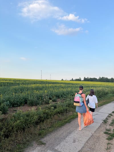 The image depicts two individuals walking along a gravel path beside a field of yellow flowers under a clear blue sky. The sun is low in the sky, casting long shadows, suggesting it is late afternoon or early evening. The weather appears to be pleasant, with a few scattered clouds.
The person in the foreground is wearing a denim skirt, a light-colored tank top, and a black baseball cap. They are carrying a colorful backpack with a checkered pattern and holding a large, bright orange plastic bag filled with items. The second individual, further ahead on the path, is wearing a white t-shirt and dark shorts.
The setting is rural, with a vast field of yellow flowers stretching to the horizon. In the distance, a line of trees forms a dark border against the sky, and a cell tower is visible against the clear horizon. The overall mood is relaxed and casual, suggesting a leisurely walk.
There is no visible text in the image. The location is identified as Mar'inskoe, Russia.