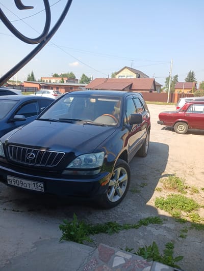 The image shows a dark blue Lexus RX SUV parked on a gravel and concrete surface. The vehicle's license plate reads "B909OT 154 RUS". To the right of the SUV, a red sedan is parked. In the background, there are houses with tiled roofs and trees. The sky is clear and blue, indicating a sunny day. The scene appears to be in a residential area, possibly a yard or a parking lot, in Prokudskoye, Russia. There are no people clearly visible in the image.