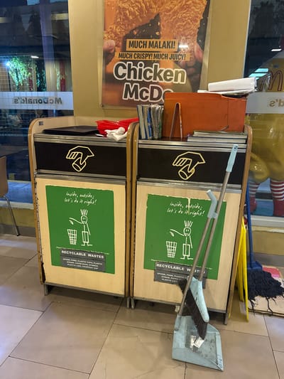 This image captures a close-up of two McDonald's recycling bins positioned side-by-side in an indoor setting, likely a restaurant. The bins have a wooden exterior with black and green panels. The green panels prominently display instructional graphics and text for recyclables. One panel shows a stylized drawing of a person throwing trash into a bin labeled "RECYCLABLE WASTES," with a list of accepted items such as spoons, forks, plastic cups, straws, and paper bags. The other bin has a graphic of a hand holding a cup, with the text "inside, outside; let's do it right!" written in a handwritten style.

To the left of the bins, a large McDonald's advertisement featuring crispy fried chicken is visible. The text on the advertisement reads "MUCH MALAKI! MUCH CRISPY! MUCH JUICY!" and "Chicken McD".

In the background, a mop with a blue handle and a blue dustpan with a brush are leaning against the bins. A bright yellow object, possibly part of a cleaning tool or signage, is also visible. A glimpse of a window reveals a blurry interior with what appears to be more McDonald's branding, including the iconic golden arches and the text "a'b" and "McDonald's" reflected. The overall lighting suggests it is daytime, and the location appears to be a well-lit fast-food establishment. No people are directly visible in the image.