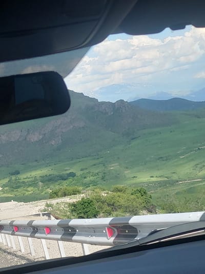 The image presents a panoramic view of a vast, mountainous landscape, likely captured from inside a vehicle traveling through Yelpin, Armenia. The scene is oriented as if rotated 90 degrees clockwise, displaying the horizon vertically.
In the foreground, elements of the vehicle frame the view. On the left, a dark dashboard, a section of the windshield, and a rearview mirror are visible. On the right, a dark vertical edge, possibly part of the car's pillar or side mirror housing, encloses the view.
The main subject is a sprawling natural environment. The sky, visible in the upper left, is bright blue with scattered white, fluffy clouds. Below the sky, layers of distant, hazy blue mountains recede into the horizon. Closer to the viewer, a prominent mountain range with rocky outcrops and abundant green foliage stands out. The landscape comprises rolling green hills and valleys, suggesting a fertile and well-vegetated area.
In the immediate foreground on the right, parallel to the vehicle, runs a distinct highway guardrail. It features a design with alternating grey and black sections and regularly spaced, reflective red markers. Beneath the guardrail, a light-colored, textured road surface or embankment is visible, along with scattered small trees and bushes.
The time of day appears to be bright daytime, under clear and sunny weather conditions. The setting suggests a journey through a scenic, mountainous region. There is no visible text in the image.