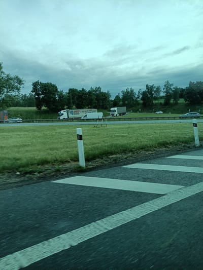 The image captures a view from a moving vehicle on a highway in Divišov, Czechia. The foreground shows a striped pedestrian crossing painted on the road, indicating a transition area. Beyond the crossing, a grassy embankment lines the highway, leading to a road with several vehicles. A white semi-trailer truck with the logo "NAD" and Slovakian text, a white panel van, and a white car are visible on the highway. Further in the background, more trees and a cloudy sky are present. The overall scene depicts a typical highway landscape under overcast conditions.