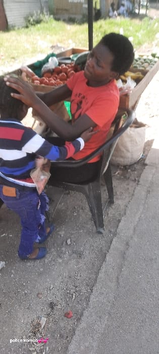 An outdoor scene in Homa Bay, Kenya, depicts an adult and a child near a produce stall. An adult individual, with dark skin and short dark hair, is seated on a black plastic chair. They wear a red short-sleeved t-shirt with a black graphic and black pants. Their right arm is around the shoulder of a child, and their left hand rests on the child's head. The adult is looking down at the child. The child, standing in front of the adult with their back partially to the camera, wears a long-sleeved striped top (red, white, blue, black), distressed blue jeans with a brown patch on the right hip, and blue sandals. The child holds a small, clear plastic bag containing reddish-brown items. To the right and behind the adult is a wooden market stall displaying various produce, including a pile of red tomatoes, green produce (possibly watermelons), and other unidentifiable items. A large, light-colored woven sack is on the ground behind the chair. The ground consists of uneven dirt and gravel, with a concrete or paved edge running vertically on the right side of the frame. A small red object lies on the ground in the foreground. The background shows green grass and distant structures under daylight conditions.