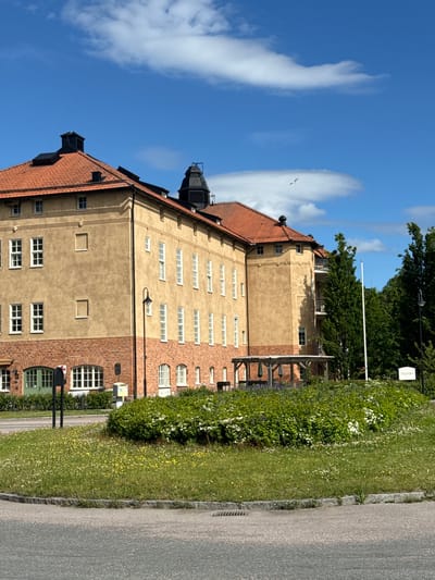 The image depicts a large, yellow building with a red tile roof. The building has multiple floors and numerous windows. The lower portion of the building is made of red brick. In front of the building, there is a round garden area with green bushes and yellow and white flowers. A paved road curves in front of the garden. A signpost with text in Swedish is visible to the right of the building. The sky is blue with scattered white clouds. The building appears to be the "Mogsjorden" building, a part of the former hospital in Eksjö, Sweden.
