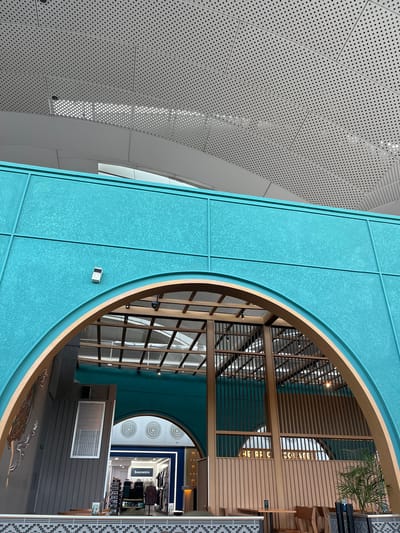 This is an interior architectural photograph, likely taken in a modern building in Ramunia Dua, Indonesia. It features a view looking through a large circular opening framed in wood and teal-colored panels.
To the left is a perforated white metal wall. The area beyond the circular opening shows a multilevel space with wooden structures, and storefronts with merchandise labeled "Souvenirs". The lighting appears to be a mix of natural and artificial.