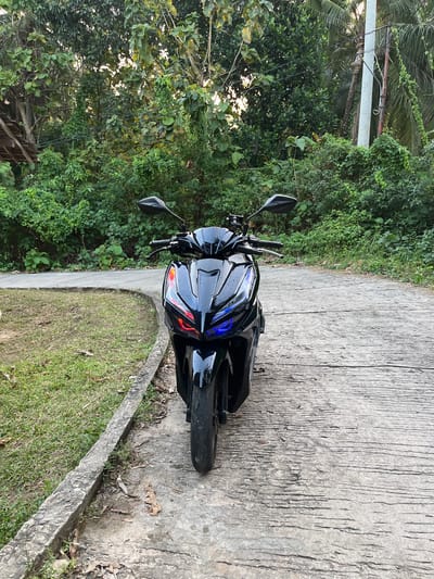 A black motorcycle is parked on a concrete path with a grassy area to the left, in Legonipah, Indonesia. The motorcycle is facing the camera and has its headlights on, emitting red and blue light. Lush green trees and vegetation surround the scene. A utility pole and wires are visible in the background. The ground is made of rough concrete with visible cracks and debris.