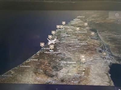 This image displays an in-flight map on an electronic screen, depicting the Eastern Mediterranean region. The map shows the coastline of Israel and parts of Lebanon, with the dark blue Mediterranean Sea on the left (west) and a detailed landmass to the right (east). Numerous cities are labeled, including Beirut, Sidon, Nahariyya, Haifa, Netanya, Tel Aviv, Rishon LeZion, Ashdod, Ashkelon, and Gaza City along the coast, and Nazareth, Nablus, Jerusalem, Jericho, Bet Shemesh, Hebron, and Irbid inland. Several generic building icons and specific airport icons (airplane symbol within a square) are placed near various locations. A white silhouette of an airplane is positioned over the Mediterranean Sea, west-southwest of Tel Aviv and west of Rishon LeZion. A smaller, light blue teardrop-shaped marker with a white cross, indicating the current position, is located just south of the aircraft icon, west of Rishon LeZion. The city of Or Yehuda, Israel, is not explicitly labeled on this map. Geographically, Or Yehuda is situated inland, east-southeast of Tel Aviv and northeast of Rishon LeZion. The displayed aircraft is located over the Mediterranean Sea, approximately west-northwest of Or Yehuda's estimated position. The screen itself shows reflections from ambient light, indicating its surface.