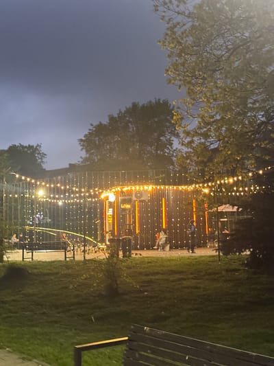 A nighttime scene depicts an outdoor area. A building or structure illuminated with orange and white lights is partially visible, adorned with multiple strings of small lights. Trees are present on the left and in the background, silhouetted against a dark sky. On the right, a section of a wooden bench is visible, with grass and possibly a pathway extending into the background. People are seated and standing in front of the illuminated building. This is Komgaron, Russia.