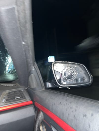 From inside a vehicle, a side mirror is missing its glass. The dark night is visible beyond the damaged mirror. The dashboard is a dark gray, and a red trim accents the vehicle's interior. Air vents are visible, as is a cloudy reflection on the driver's side window. The image is blurry, suggesting motion.