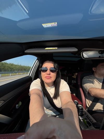 A woman is driving a car on a sunny day. She is wearing sunglasses and a light-colored t-shirt. The interior of the car is visible, including the dashboard, seats, and a panoramic sunroof. Outside the car, trees and a highway are visible. There are two cans of soda in the center console of the car, and a man is in the passenger seat. A child is visible in the back seat. The image appears to have been taken in the city of Fyodorovo, Russia.