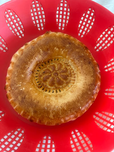 A close-up, top-down view shows a freshly baked, golden-brown flatbread with a patterned center and sesame seeds sprinkled on top. The bread is resting inside a bright red plastic basket with a distinctive criss-cross pattern in its oval-shaped openings, a common sight in markets and bakeries. The background is blurred, suggesting the photo was taken in a market environment in the city of Kara-Suu, Kyrgyzstan, where such bread is a staple. The lighting is bright and natural, highlighting the texture and color of the bread.