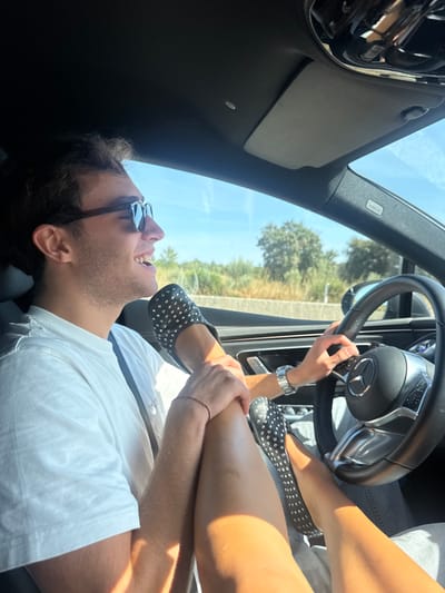 The image depicts the interior of a modern vehicle on a clear, sunny day. A male driver, visible from the chest up, occupies the driver's seat. He has dark, curly hair, wears dark sunglasses, a white short-sleeved t-shirt, and a thin red string bracelet on his left wrist. He is smiling with his mouth open, looking towards the right side of the frame. His right hand grips the steering wheel, which features a Mercedes-Benz logo. A silver watch is visible on his right wrist.
Across the driver's lap and thighs are the bare legs of a second individual, not otherwise visible. Both feet of this individual wear black flat shoes adorned with silver studs. One foot rests on or near the dashboard area, while the other is positioned close to the steering wheel. The male driver's left forearm and hand rest on the thigh of the second individual.
Through the vehicle's windows, a bright blue sky is visible above a landscape dominated by green foliage, primarily trees and bushes, suggesting a rural or semi-rural setting. A light-colored concrete barrier is discernible horizontally in the background, indicative of a roadside environment. While the request specifies Alcanena, Portugal, no specific visual landmarks or identifiers within the image itself confirm this precise geographic location.