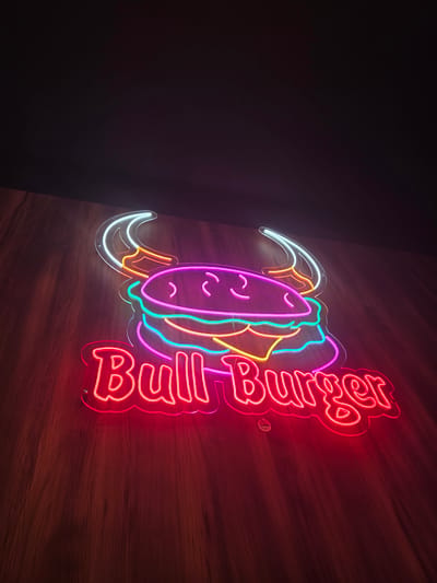The image shows a neon sign for "Bull Burger" mounted on a wall with a wood-like texture. The sign is multi-colored, with red outlining the words "Bull Burger" and other colors highlighting the burger image to the left. The burger image has a pink bun with a smiley face, white horns, orange cheese and lettuce and green meat. The photo is taken indoors and the left portion of the image is dark, suggesting a dimly lit environment. The location in the city of Dolni Chiflik, Bulgaria is not clear from the image.
