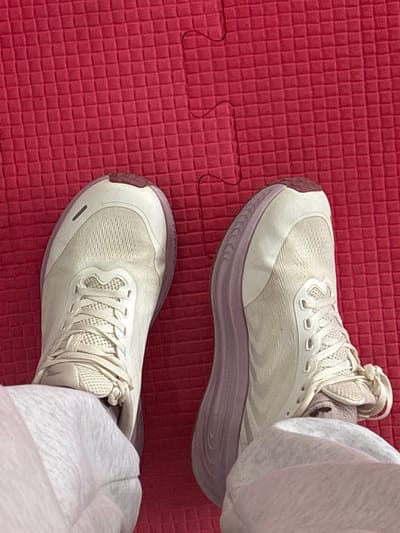 The scene is situated in Tayshet, Russia, within an indoor environment. The image presents a downward-facing perspective of an individual's lower legs and feet resting on a red textured foam puzzle mat. The subject is wearing light-colored, possibly off-white or light grey, athletic trousers that obscure the ankles and partially cover the upper sections of the shoes. Two athletic shoes, primarily off-white or very light beige in color, are visible. These shoes feature pale lavender or light purple soles/midsoles, with a small reddish-brown segment on the toe and heel areas of the outsole. The uppers are constructed from a mesh-like material, and the laces are tied. The shoes exhibit some signs of minor wear. The underlying surface is a bright red foam mat composed of interlocking puzzle pieces, characteristic of a gym floor or activity area. The mat's surface shows a fine, square grid pattern. Two distinct mat pieces are visible, with their wavy interlocking seam running from the lower right to the upper central portion of the frame. No other objects or individuals are discernible in the immediate vicinity.