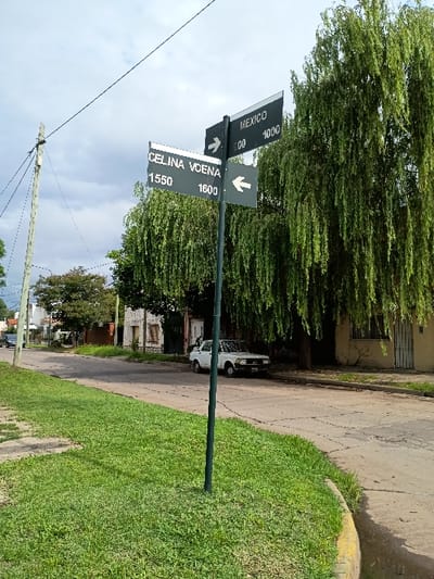 The image shows a street corner in El Talar, Argentina, located at coordinates -34.467, -58.644. A street sign post with two signs points to the left and right. The left sign is for “Celina Voena” with the numbers 1550 and 1600. The right sign is for “Mexico” with the numbers 1000 and 00. A white car is parked on the right side of the street. There is a tree with green leaves on the right side of the image. The ground is paved and there is a patch of green grass to the left. The sky is overcast.