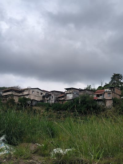 The image depicts a cluster of multi-story buildings situated on a hillside in the city of Payatumpi Baru, Indonesia. The buildings, some appearing to be older and in varying states of repair, are nestled amongst lush green vegetation. The sky above is overcast with dark, heavy clouds, suggesting impending rain or stormy weather. In the foreground, tall grasses and some discarded white plastic bags are visible, indicating a somewhat unkempt area at the base of the hillside. The architecture of the buildings varies, with some featuring balconies, satellite dishes, and visible windows. The overall scene conveys a sense of a densely populated hillside settlement under a dramatic sky.