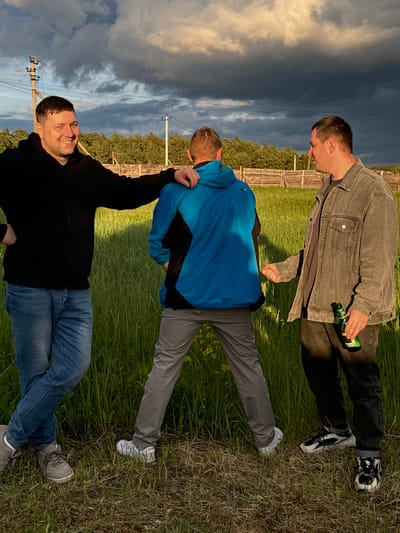 The image shows three men in a field in Vysokinichi, Russia. The man at the top wears a gray jacket, black pants, and holds a green bottle. The middle man wears a blue jacket and gray pants. The bottom man wears a black jacket and blue jeans and has his arm raised. The background includes a cloudy sky, trees, and power lines. A wooden fence is visible between the top and middle men. The men are positioned vertically in the frame and are lying down.