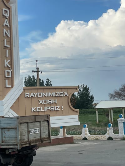 The image shows a street scene in Imeni Andreyeva, Uzbekistan. A sign with Cyrillic and Latin letters reads "RAYONIMIZGA XOSH KELIPSIZ!", which translates to "Welcome to our district!". Below, another sign shows letters that appear to be "O'ZBEKISTON" and "DANZI-KOL". A truck with a license plate "95 S 923 TA" is parked to the right of the sign. The sky is cloudy, with a mix of blue sky and white clouds. The weather appears to be overcast. The scene is set in a town or city, with buildings and trees visible in the background. The overall mood is welcoming and informative.