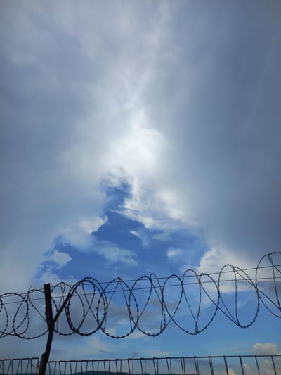 The image shows a barbed wire fence against a cloudy sky. The sky is a mix of white clouds and patches of blue sky, with sunlight breaking through in the center. The barbed wire fence is prominent in the foreground, with several coils of wire attached to a metal post. The lower portion of the image shows the top of another fence, with hills visible in the distance. The image is taken from a low angle, looking upwards. There are no people or animals visible in the image. The scene is likely outdoors, possibly in a rural or industrial area.