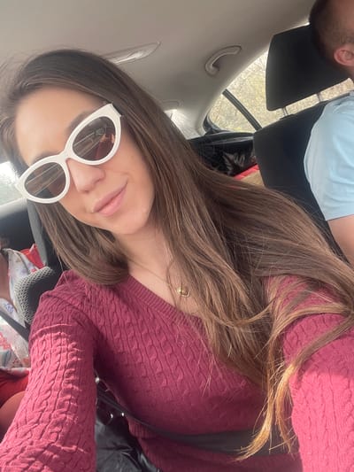 A selfie taken inside a car. A fair-skinned woman with light brown hair and white framed sunglasses is wearing a maroon cable knit sweater. The car appears to be parked, indicated by the headrest in view and the outdoor scenery visible through the rear window. The location, Kiosevtsi, Bulgaria, is not discernable from the visible scene.