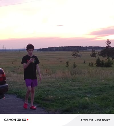 A young male, appearing to be in his late teens or early twenties, stands on a paved surface in a rural outdoor setting. He is wearing a black t-shirt with colorful graphic print and purple shorts. His feet are clad in red crocs. The male is holding a smartphone in his right hand, looking down at it. To his left, the rear section of a dark-colored vehicle is visible. The background consists of a vast, grassy field with scattered trees and power lines extending into the distance. The sky above is a blend of light yellow and pink hues, suggesting either sunrise or sunset. The overall scene appears to be located in Алтынды ауылдық округі, Kazakhstan, given the context.