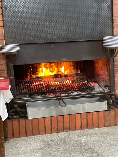 This image depicts an active, large-scale, wood-fired grilling station, likely situated within a restaurant in Mogadouro, Portugal. The primary structure is built from red-brown bricks, forming a sturdy base and a wide fireplace opening. Above the firebox, a dark, textured diamond plate metal panel extends upwards, framed by black metal.
Within the fireplace opening, a vibrant wood fire burns, emitting bright orange and yellow flames and illuminating glowing red embers below. A black metal grill grate is positioned over these embers, and several pieces of dark, seared meat are visible cooking on its surface. Directly below the front edge of the grill, a long, rectangular stainless steel drip tray is in place, with a pair of metal tongs resting within it.
To the left and right of the central grilling opening, black metal fixtures resembling wall sconces are mounted, each supporting a stack of approximately ten white, rimmed plates. On the far left, a portion of a wooden table with a white tablecloth and a red object (possibly a menu holder or napkin dispenser) is partially visible. The floor in the immediate foreground consists of light grey, possibly tiled, surface. No individuals are present in the frame.