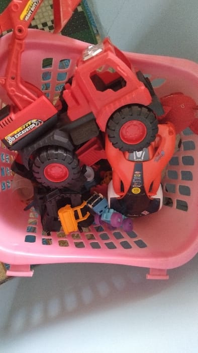 A pink plastic basket with a grid pattern sits on a white surface, containing several plastic toy vehicles.  The basket is slightly tilted, and the toys are piled in a haphazard manner.  The main toy is a red toy excavator with a black cab and black wheels.  The excavator has a sticker on the side that reads "DG Excavator".  Behind the excavator are other toys including a red toy car with black and blue accents, and a yellow and blue toy truck. The toys are all in focus and appear to be new.  The scene is located in Kecamatan Panjalu, Indonesia, at latitude -7.143, longitude 108.278.