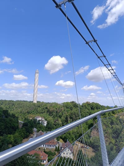 The image shows a view from a suspension bridge in the city of Rottweil, Germany. In the foreground, a metal railing and mesh fence are visible, suggesting the perspective is from the bridge's walkway. The bridge structure with its cables and supports is also prominent. In the middle ground, lush green trees cover rolling hills, and several buildings with red-tiled roofs are nestled amongst the foliage. Further in the background, a distinctive, tall, white tower with a spiraling design stands out against a bright blue sky dotted with white clouds. A railway track is also visible winding through the landscape. The overall scene depicts a scenic view of Rottweil, with the prominent tower likely being the test tower.