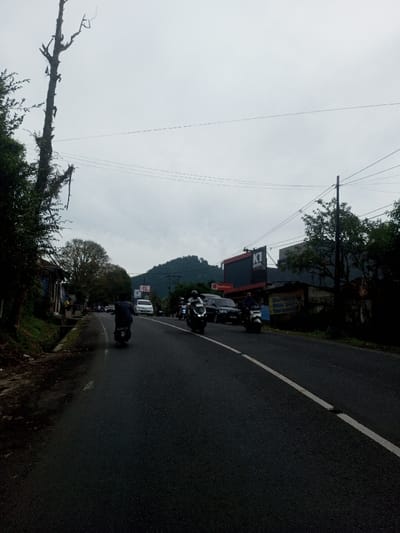 The scene depicts a moderately busy road in Sat Thay (East), Myanmar, under an overcast sky. The asphalt road surface appears dark, possibly damp, and features a solid white line marking the shoulder on the right and a broken white center line. Multiple vehicles are in motion: a person on a dark scooter, wearing a plaid shirt, proceeds away from the viewer in the foreground-left lane. Further down, another individual on a dark scooter also moves away. Approaching from the mid-ground in the right lane are two individuals on scooters, both wearing helmets. Several cars, including a white sedan and a dark SUV, are visible further down the road, some parked or moving slowly.
On the left side of the road, a bare, gnarled tree trunk with sparse branches extends vertically into the sky, partially obscured by dense green foliage lining the roadside. A drainage ditch runs parallel to the road on this side, next to a series of indistinct low-rise structures.
The right side of the road features a red commercial building with a prominent black sign displaying "K1 HOTEL" and "RedDoorz" branding. Adjacent to this, a smaller, weathered structure with a blue tarp roof and a partially legible white banner is visible. A dark utility pole stands on the right, supporting multiple overhead power lines that traverse the upper portion of the frame. In the background, a large, dark green mountain range rises beneath the cloudy sky, suggesting a natural landscape surrounds the urban elements.