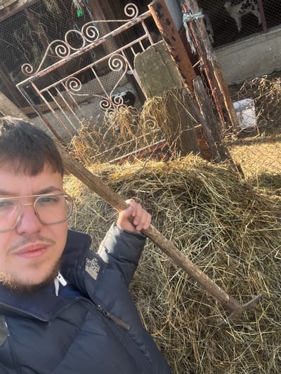 The image depicts a male individual in a selfie-style shot in an outdoor, rural setting identified as Dzhurovo, Bulgaria, during daylight hours. The man, with short dark hair, light facial stubble, and wearing gold-rimmed rectangular eyeglasses, is dressed in a dark blue or black puffer jacket with "THE NORTH POLE" branding visible on the left sleeve. He holds a wooden-handled hayfork with a three or four-tined metal head, resting it against his right shoulder. Behind the man, a large pile of dry hay or straw is present. In the background, an animal enclosure is visible, constructed from wire mesh fencing, weathered wooden posts, and rusted metal supports. Within this enclosure, an ornate, rusted metal gate featuring scrolled designs is positioned. Several small animals, identified as lambs or young goats, with black and white spotted coats, are present inside the enclosure. A white bucket or container is visible behind the fence on the right side of the frame.