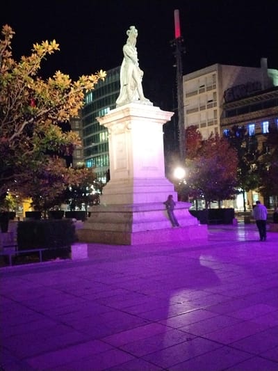 The image depicts the Place de Jaude in Clermont-Ferrand, France, at night. The central feature is the light-colored statue of Vercingétorix, a standing male figure with a crown, positioned on a multi-tiered stone pedestal. The ground of the urban square is intensely illuminated by a purple light, reflecting off the large square pavement tiles and the lower sections of the statue's base.
One individual, dressed in dark clothing, is seated on the lower steps of the pedestal, casting a discernible shadow on the purple-lit ground. Further to the right, a second person, wearing a light-colored top and dark trousers, is observed walking away from the statue.
In the background, a tree with yellowish-brown autumn foliage is partially visible to the left of the statue, in front of a modern building featuring multiple illuminated windows. To the right of the statue, a tall, slender structure topped with a red light stands against the dark sky. Beyond it, a multi-story building with a light-colored facade is visible, adjacent to another building displaying distinct blue-lit windows. The sky is dark, indicating a nighttime setting.